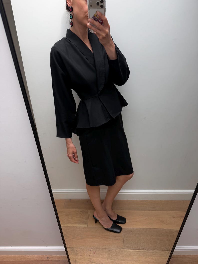 Puede incluir: Un conjunto negro de dos piezas compuesto por una chaqueta de manga larga con peplum y cuello chal, y una falda a juego a la altura de la rodilla. La chaqueta presenta una cintura entallada y un bajo acampanado, junto a una falda recta y mules negros.