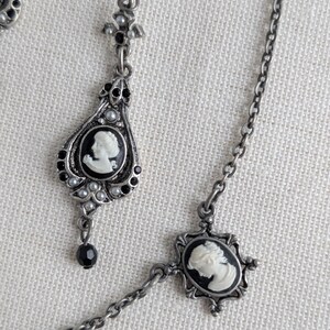 May include: Antique silver-toned jewelry set featuring cameo pendants. The set includes a pair of teardrop earrings and a necklace. Each piece has a black and white cameo design, accented with small pearls and black beads. The jewelry has an ornate, vintage aesthetic.