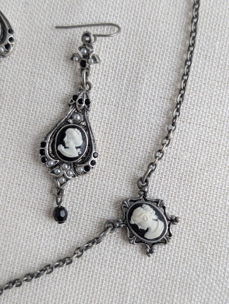 May include: Antique silver-toned jewelry set featuring cameo pendants. The set includes a pair of teardrop earrings and a necklace. Each piece has a black and white cameo design, accented with small pearls and black beads. The jewelry has an ornate, vintage aesthetic.