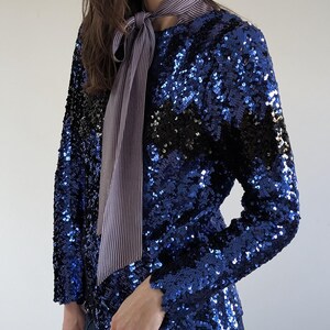 May include: A long-sleeved, sequined top in shades of blue and black. The top features a striped necktie in shades of grey and white. The sleeves have a scalloped edge. The top is paired with blue jeans.