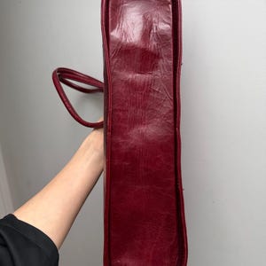 May include: A burgundy tote bag with a rectangular shape and rounded corners. The bag has a textured surface and two thin handles. The bag is held vertically, showcasing its side profile.