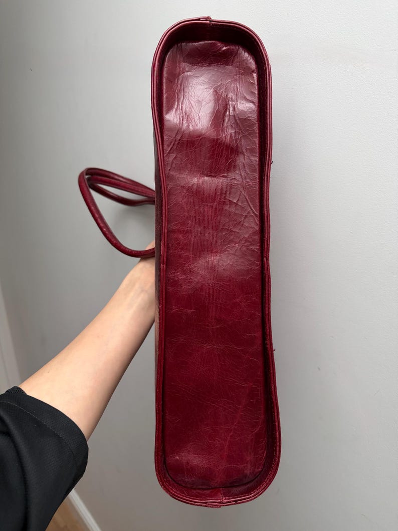 May include: A burgundy tote bag with a rectangular shape and rounded corners. The bag has a textured surface and two thin handles. The bag is held vertically, showcasing its side profile.