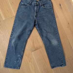 Op de afbeelding: Blauwe spijkerbroek met een klassiek ontwerp met vijf zakken. De jeans heeft een rechte pijp en vertoont wat slijtage, waaronder een vlek op het linkerdeel. De jeans liggen plat op een houten oppervlak.