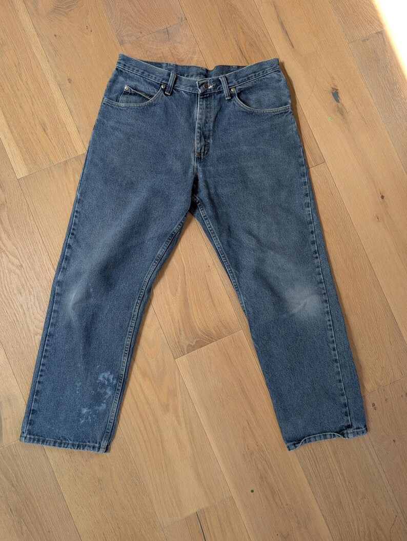 Op de afbeelding: Blauwe spijkerbroek met een klassiek ontwerp met vijf zakken. De jeans heeft een rechte pijp en vertoont wat slijtage, waaronder een vlek op het linkerdeel. De jeans liggen plat op een houten oppervlak.