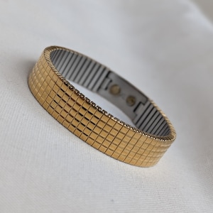 May include: A gold-toned expandable bracelet with a silver metal band. The bracelet is made of small square links.