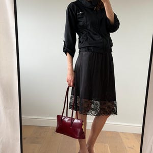 May include: A black jacket with a high collar and a black skirt with a lace hem. A burgundy leather handbag with long handles is held in the hand. Black slingback heels complete the outfit.