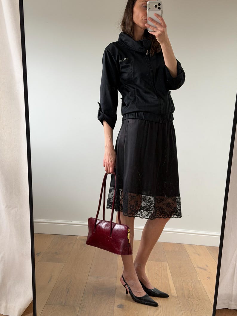May include: A black jacket with a high collar and a black skirt with a lace hem. A burgundy leather handbag with long handles is held in the hand. Black slingback heels complete the outfit.