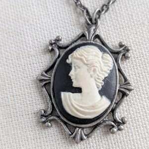May include: A silver-toned necklace featuring a cameo pendant. The pendant has a black background with a white profile of a woman. The cameo is set in an ornate, decorative frame.
