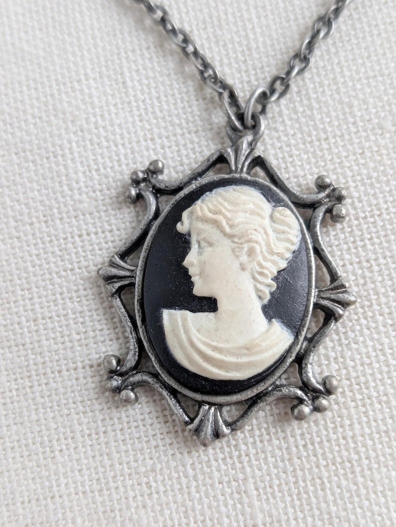 May include: A silver-toned necklace featuring a cameo pendant. The pendant has a black background with a white profile of a woman. The cameo is set in an ornate, decorative frame.