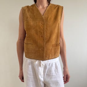 May include: A tan suede vest with a V-neck, three buttons, and two front pockets. The vest has a braided trim along the edges. The model is wearing white linen pants.
