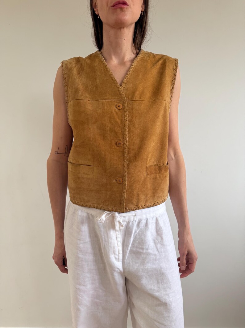 May include: A tan suede vest with a V-neck, three buttons, and two front pockets. The vest has a braided trim along the edges. The model is wearing white linen pants.