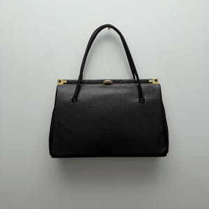 May include: A black handbag with a rectangular shape and a top handle. The bag features gold-coloured accents on the clasp and handle attachments. The bag is made of a smooth, possibly leather, material.