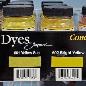 Peut inclure: Un présentoir de colorants acides dans des flacons en verre avec des bouchons noirs. Les flacons sont étiquetés avec la couleur et le numéro du colorant, notamment 600 Ecru, 601 Jaune Soleil, 602 Jaune vif et 603 Jaune doré. L'étiquette indique également "Concentré, adapté à l'utilisation sur la soie, le nylon et autres fibres protéiques."