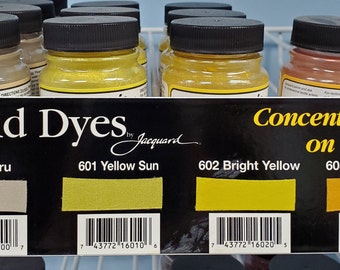 May include: A display of acid dyes in glass bottles with black lids. The bottles are labeled with the dye color and number, including 600 Ecru, 601 Yellow Sun, 602 Bright Yellow, and 603 Golden Yellow. The label also states "Concentrated, suitable for use on silk, nylon, and other protein fibers."