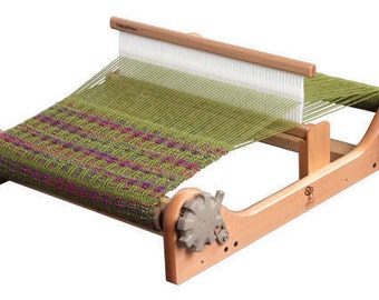 May include: A wooden loom with a green and purple woven fabric on it. The loom has two wooden spools on either end and a wooden shuttle in the middle. The loom is made by Schacht.