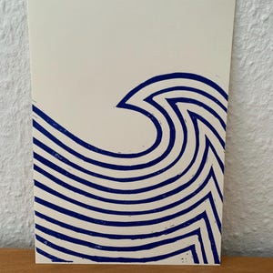 Könnte beinhalten: Ein cremefarbener Kunstdruck mit einem stilisierten Wellenmuster in Marineblau. Die Welle besteht aus gebogenen, parallelen Linien, die ein Gefühl von Bewegung erzeugen. Der Druck befindet sich auf einem rechteckigen Papier, mit einer minimalistischen Ästhetik.