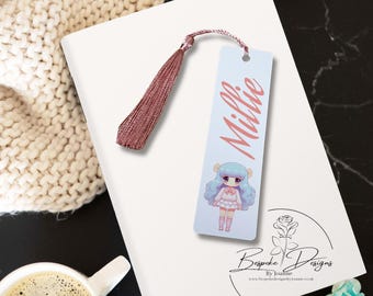 May include: A white bookmark with a pink tassel, featuring a cartoon girl with blue hair and a pink dress. The bookmark has the name "Millie" written in pink on it. The bookmark is on a white surface with a cup of coffee and a small jar of candles.