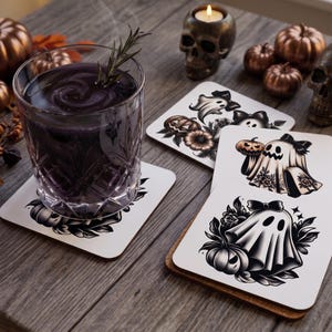 May include: A set of square drink coasters featuring black and white illustrations of ghosts wearing bows, surrounded by pumpkins and floral elements. One coaster supports a glass filled with a dark purple liquid and a sprig of rosemary.