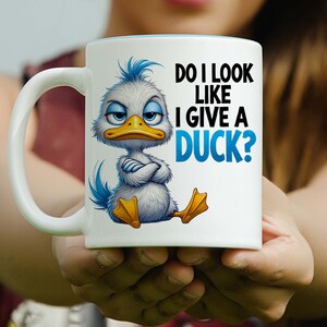 May include: White ceramic mug with a blue interior and a handle. The mug features a cartoon duck with crossed arms and the text "DO I LOOK LIKE I GIVE A DUCK?" in black and blue.