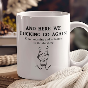 May include: White ceramic mug with a black handle. The mug features the text "AND HERE WE FUCKING GO AGAIN" and "Good morning and welcome to the shitshow" with a stick figure illustration. The mug is on a wooden surface.