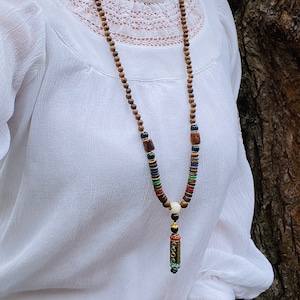 May include: A brown wooden beaded necklace with colorful accents and a small gold pendant. The necklace is worn by a person wearing a white blouse.