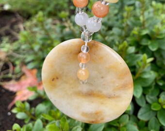 May include: A round, light brown pendant with a white and brown marbled pattern hangs from a multi-colored beaded necklace. The necklace features white, light green, orange, and light brown beads.