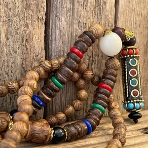 May include: A wooden beaded necklace with colorful accents. The necklace features a white bead, a long wooden cylinder with colorful accents, and black beads.