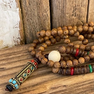 May include: A wooden prayer bead necklace with a decorative pendant featuring turquoise, red, and gold accents. The necklace is made of brown wood beads and includes a white bead.