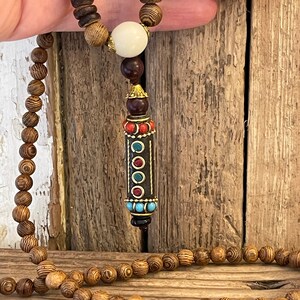 May include: A wooden beaded necklace with a decorative pendant. The pendant is made of dark wood with gold accents and features red, blue, and turquoise stones.