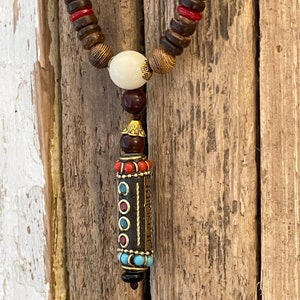 May include: A wooden beaded necklace with a decorative pendant. The pendant is a small cylinder with a gold-colored metal top and bottom. The cylinder is decorated with red, blue, and turquoise stones.