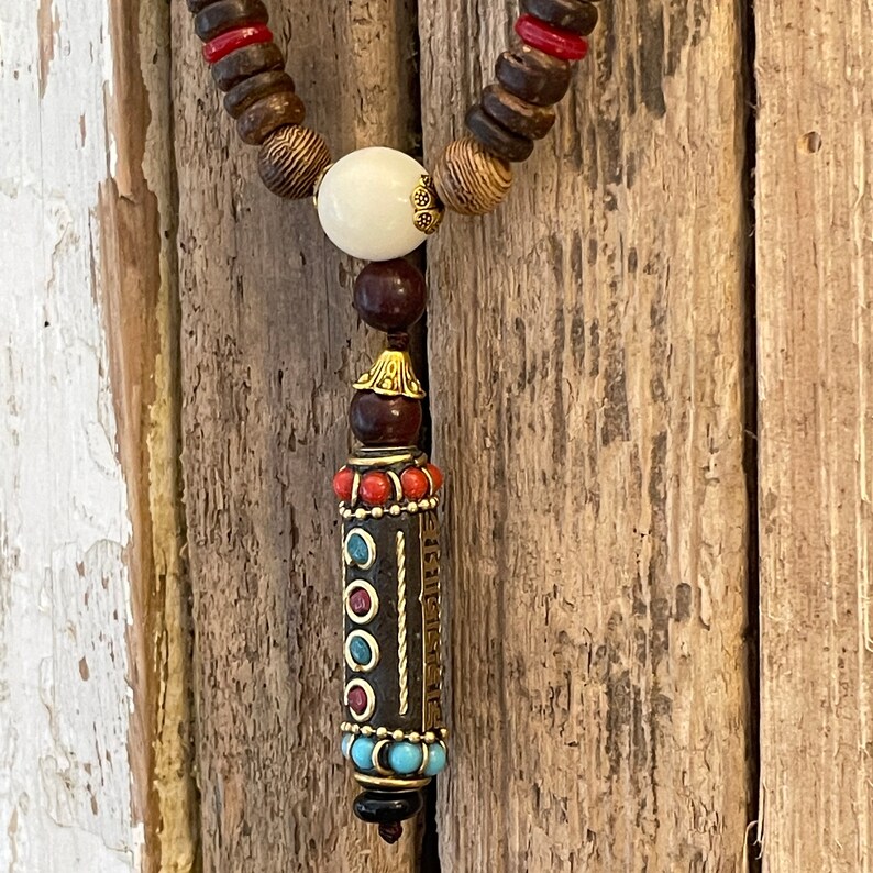 May include: A wooden beaded necklace with a decorative pendant. The pendant is a small cylinder with a gold-colored metal top and bottom. The cylinder is decorated with red, blue, and turquoise stones.