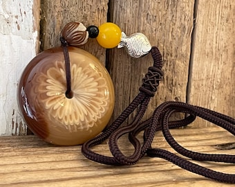 May include: A brown and beige round pendant with a floral design hangs from a brown cord with a silver fish charm and yellow and brown beads.