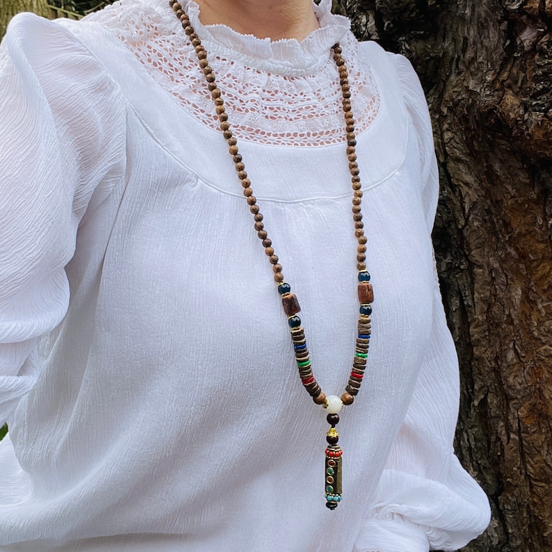 May include: A brown wooden beaded necklace with colorful accents and a small gold pendant. The necklace is worn by a person wearing a white blouse.