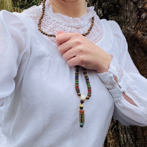 May include: A wooden beaded necklace with a colorful, multi-colored, and patterned pendant. The necklace is worn by a person wearing a white blouse with a lace collar.