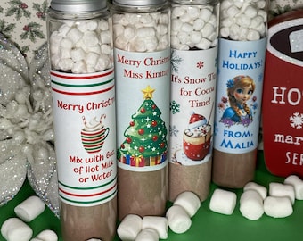 May include: Four clear glass tubes with silver screw caps filled with brown cocoa powder and white mini marshmallows. Each tube features a unique holiday-themed label with text including Merry Christmas, It's Snow Time, and Happy Holidays.