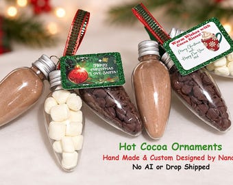 May include: Clear plastic light bulb-shaped containers filled with hot cocoa mix, chocolate chips, and white marshmallows. Each ornament features a silver screw cap, a green glittery tag with holiday text, and a red and green ribbon hanger.
