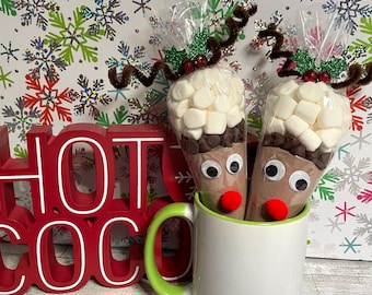 May include: Two clear plastic cones filled with hot cocoa mix, chocolate chips, and white marshmallows, decorated with googly eyes, red pom-pom noses, and brown pipe cleaner antlers, sitting in a white mug next to a red sign reading HOT COCO.