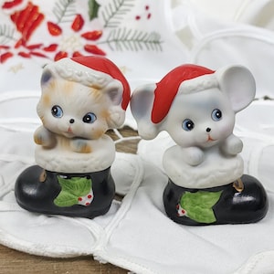 May include: Two ceramic Christmas figurines: a kitten and a mouse, each wearing a Santa hat and sitting in a black boot with a green leaf and red berry design. The kitten is cream and tan, and the mouse is white.
