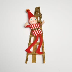 Peut inclure: Ornement de Noël vintage représentant un lutin en feutre, vêtu d'une tenue rayée rouge et blanche, grimpant une échelle en bois peinte en or. Le lutin porte un chapeau rouge avec un pompon et une expression surprise. Décoration de vacances.
