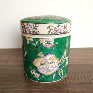 Puede incluir: Un recipiente cilíndrico de cerámica con tapa, con una base verde vibrante adornada con patrones florales en rosa, amarillo y blanco. La tapa y el borde superior tienen una banda de color crema con más diseños florales. Para almacenamiento.