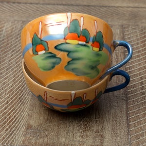 Op de afbeelding: Twee gestapelde theekopjes met blauwe handvatten. De kopjes zijn versierd met een landschapsscène met groene bomen, oranje huizen en een blauwe rivier. De kopjes zijn gemaakt van porselein en hebben een glanzende afwerking.