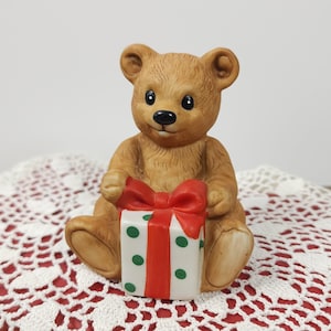 May include: A brown ceramic teddy bear figurine holding a white gift box with green polka dots and a red bow. The bear has a black nose and is sitting down. The figurine is on a red and white lace surface.