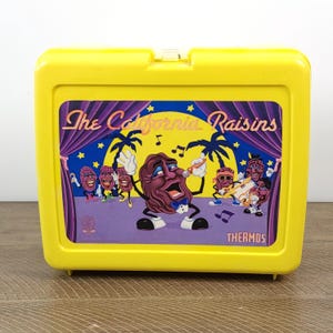 May include: A bright yellow lunchbox featuring a graphic of The California Raisins band performing on stage. The image includes the band members, musical notes, and the words "The California Raisins" and "Thermos".
