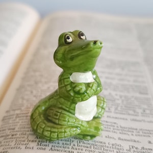 May include: A small, green ceramic alligator figurine with a white chest and a detailed, textured body. The alligator is sitting upright with its arms crossed, and its eyes are looking upwards. The figurine is placed on an open book.