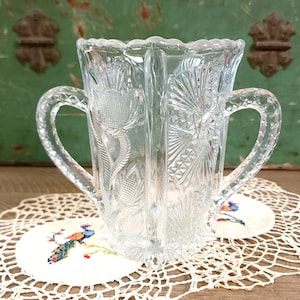 May include: Clear, vintage glass pitcher with two handles. The pitcher features intricate floral and geometric patterns. The handles are curved and attached to the sides. The pitcher is sitting on a white doily with a colorful embroidered peacock design.