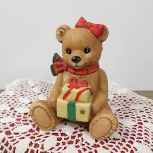 May include: A brown teddy bear figurine with a red bow on its head and a red and green scarf. The bear holds two yellow gift boxes tied with red ribbons and a green ribbon. The figurine is sitting on a white and red lace doily.