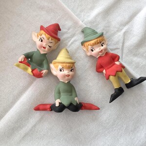 以下が含まれることがあります： 尖った帽子と耳を持つ3体の小さなエルフのフィギュア。1体は赤い帽子と緑のチュニック、もう1体は黄色い帽子と緑のチュニック、3体目は緑の帽子に赤いチュニックと黄色いズボンを着用しています。それぞれに塗装された顔の特徴があります。