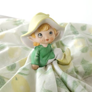 May include: A ceramic elf figurine with a yellow hat and green top, seated on a lemon-patterned cloth. The elf has blonde hair, rosy cheeks, and a whimsical design. The cloth is white with a repeating pattern of green lemons.