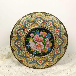Op de afbeelding: Een ronde metalen trommel met een centraal donkerblauw cirkelvormig paneel versierd met een levendig boeket van roze lelies, kleine blauwe bloemen en gele accenten. De buitenrand toont een ingewikkeld, kleurrijk en herhalend bloemmotief.