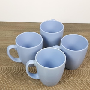 Puede incluir: Cuatro tazas de cerámica azul claro con asas curvas dispuestas sobre una superficie de madera. Las tazas tienen un acabado liso y brillante y un diseño sencillo. El interior de las tazas también es azul claro.